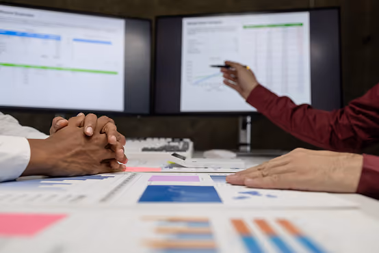 Two people discussing charts and graphs on paper with data displayed on dual monitors in background.