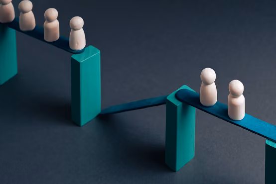 Wooden figures balancing on teal planks supported by blocks on a dark surface.
