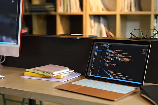 Laptop displaying code on desk with notebooks and glasses in an office with bookshelves.