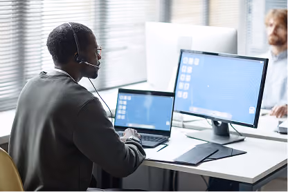 Man wearing headset working on laptop and monitor in a bright office with blinds and coworker nearby.