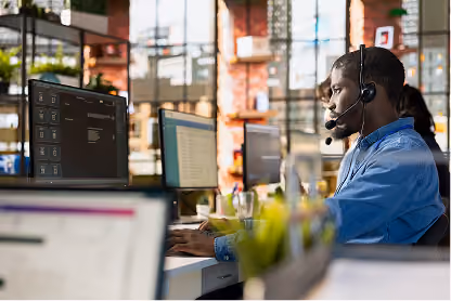 Man wearing headset working on computer in modern office with large windows and exposed brick walls.