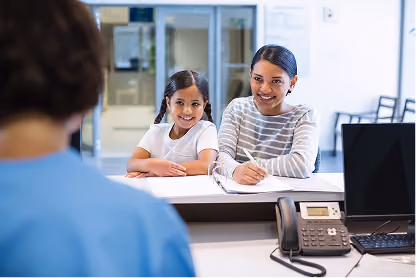 Smiling woman and girl sitting at a desk speaking with a healthcare worker across a reception window.