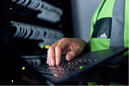 Person in green safety vest typing on a keyboard near server racks with blinking lights.