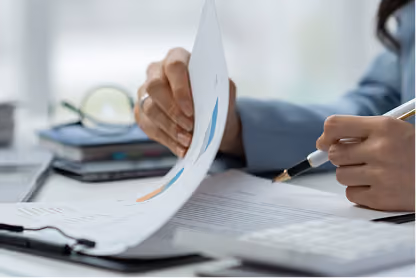 Person flipping through papers while holding a pen at a desk with documents and glasses.