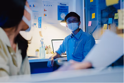 Two masked coworkers having a discussion in an office with charts and sanitizer visible.