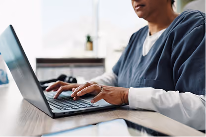 Person typing on a laptop keyboard at a wooden desk near a window with blurred background.