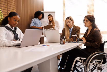 Diverse team in an office: two women at laptops, one woman in wheelchair using smartphone, and two colleagues discussing.