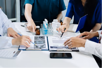 Medical professionals in scrubs discussing scans and documents around a white table with medicine bottles.
