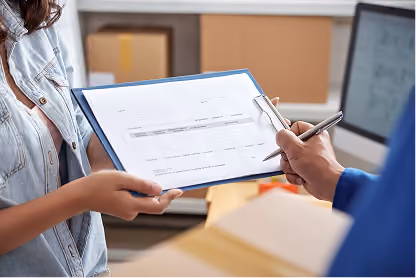 Person holding clipboard with form while another person points at it with a pen, near a computer screen.