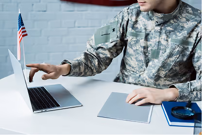 Person in military uniform pointing at a laptop on a desk with a small American flag.