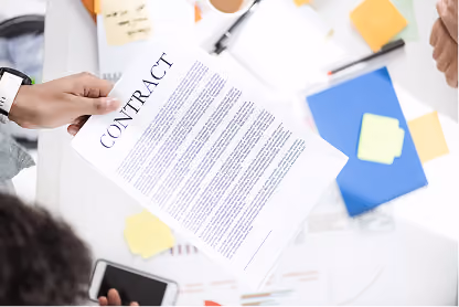 Person holding a contract document over a cluttered white desk with office supplies and sticky notes.