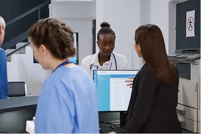 Healthcare staff and a woman in black converse in a hospital reception area with a computer screen displaying patient data.