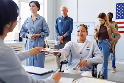 A person in a wheelchair hands a ballot to a poll worker inside a voting location.