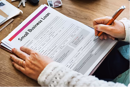 Person filling out a small business loan application form with a pen on a wooden table.