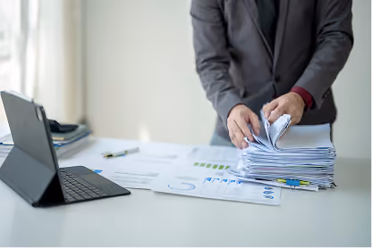Person organizing a stack of papers on a desk with documents and a tablet nearby.