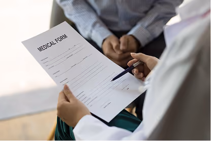 Medical professional holding a medical form and pen, patient sits across with hands clasped.