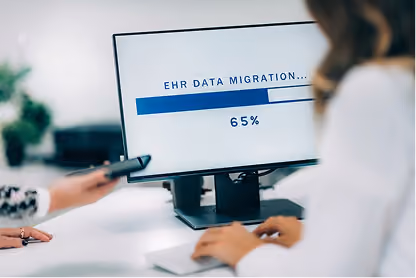 Computer screen showing EHR data migration progress at 65 percent, with two people nearby.