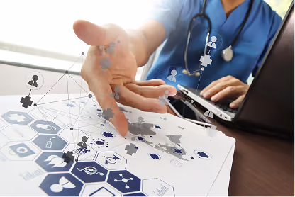 Healthcare professional in scrubs using laptop and pointing at medical data on paper with digital icons overlay.