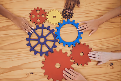 Diverse hands holding colorful wooden gears arranged on a wooden table.