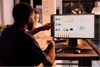 Man analyzing data charts on a computer monitor while holding a document, woman blurred in background.