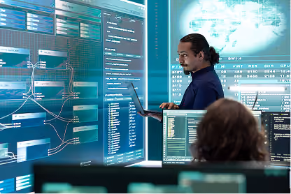 Man working on a laptop in a control room with multiple digital screens displaying data and graphs.