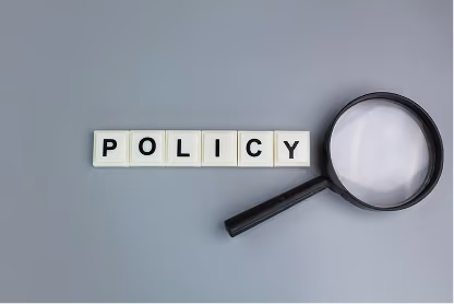 Scrabble tiles spelling POLICY next to a magnifying glass on a gray surface.