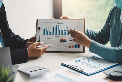 Two people analyzing business charts and graphs on paper and clipboard at a white desk.