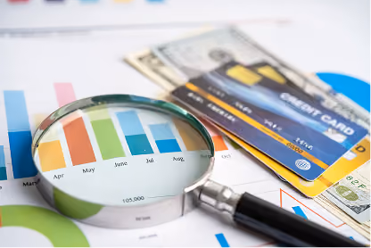 Magnifying glass over colorful bar chart with credit cards and dollar bills on financial documents.
