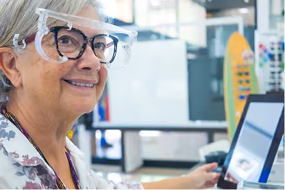 Smiling elderly woman wearing safety glasses and looking at a computer screen in a bright office.