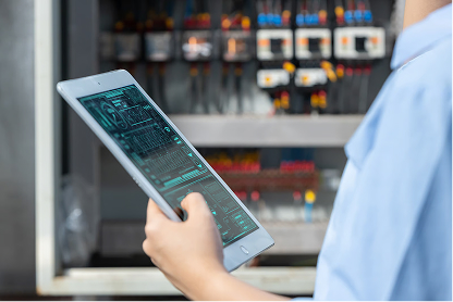 Person in blue shirt holding tablet with circuit board image near electrical control panel.