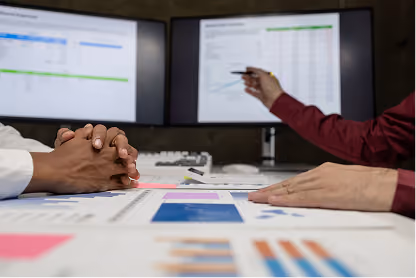 Two people discussing data with charts and graphs on desk and on dual monitors in background.
