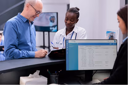 Doctor consulting a smiling older man at a clinic reception with another staff member at a computer.