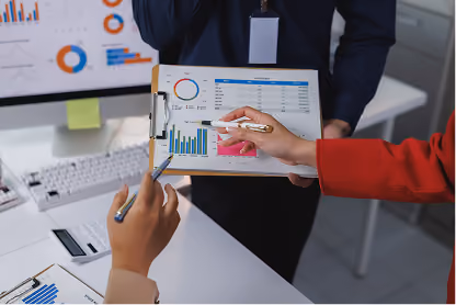 Two people in office pointing at clipboard with charts and graphs near a computer screen.