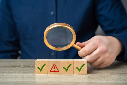 Hand holding magnifying glass over wooden blocks with green checkmarks and a red warning symbol.