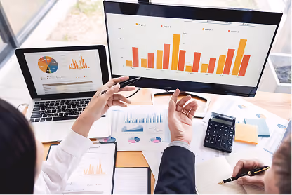 Two people analyzing colorful bar and pie charts on a laptop and monitor with documents and calculator nearby.
