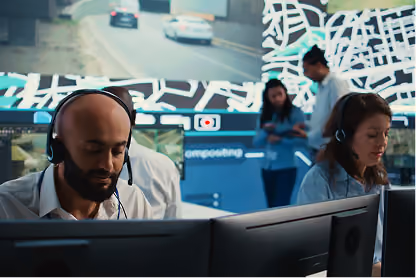 Two call center workers with headsets at desks in front of computer monitors with blurred colleagues behind.
