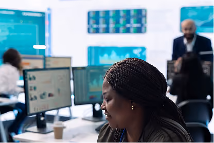 Woman working at desk with multiple computer monitors in a busy office environment.
