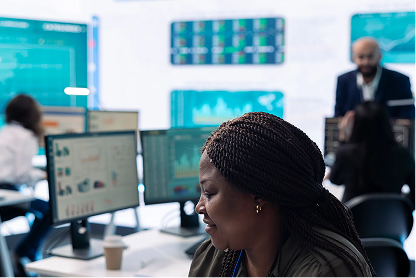 Woman working at desk with multiple computer monitors in a busy office environment.