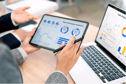 Person holding tablet showing sales performance dashboard with charts and graphs beside laptop screen.