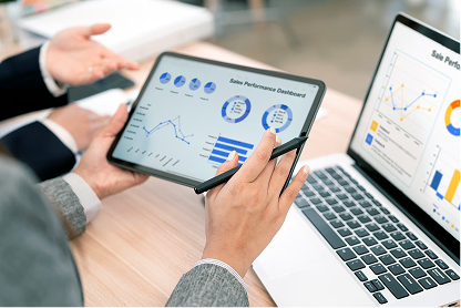 Person holding tablet showing sales performance dashboard with charts and graphs beside laptop screen.