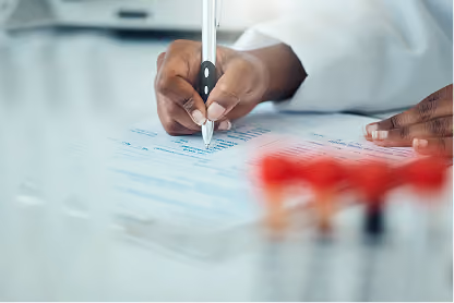 Close-up of a person’s hand writing on a medical form with lab test tubes blurred in the foreground.