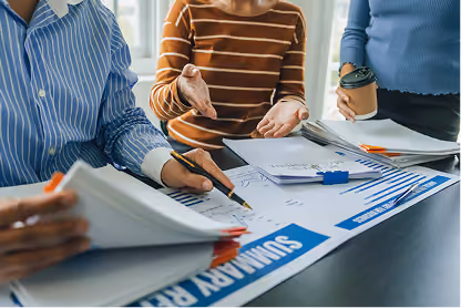 Three people reviewing summary report with charts and documents at a table, one holding a coffee cup.