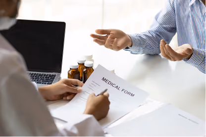 Healthcare professional filling out a medical form while a patient gestures across a desk with medicine bottles nearby.