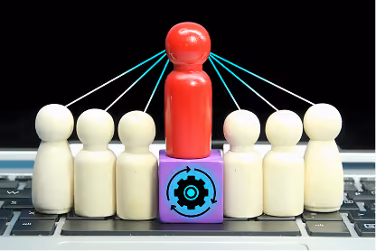 Red wooden figure on gear cube linked by strings to six white figures on laptop keyboard.
