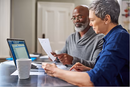 Older man and woman reviewing documents together at a table with a laptop and coffee mug.