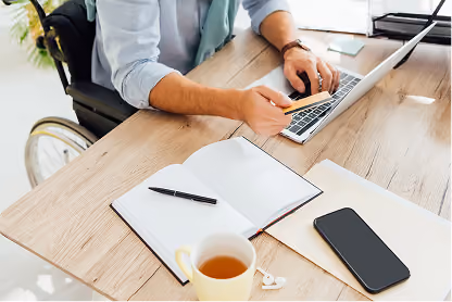 Person in wheelchair typing on laptop at desk with open notebook, smartphone, tea cup, and earphones.