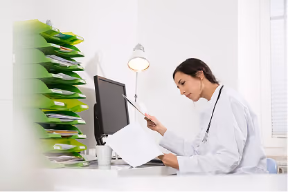 Female doctor reviewing documents at desk with computer and green file folders stacked nearby.