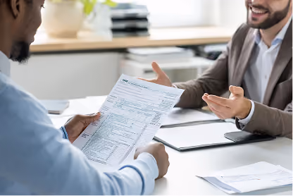 Two men discussing documents across a desk in a bright office setting.