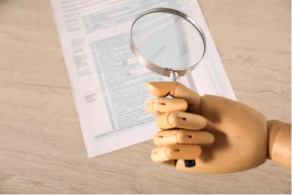 Wooden mannequin hand holding magnifying glass over a document on a wooden surface.