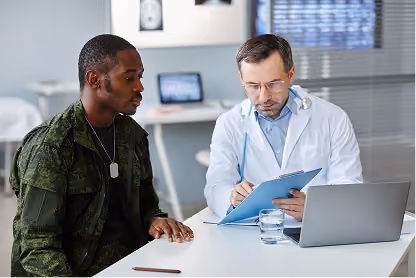 Doctor in white coat consulting a male soldier at a desk with a laptop and medical files.